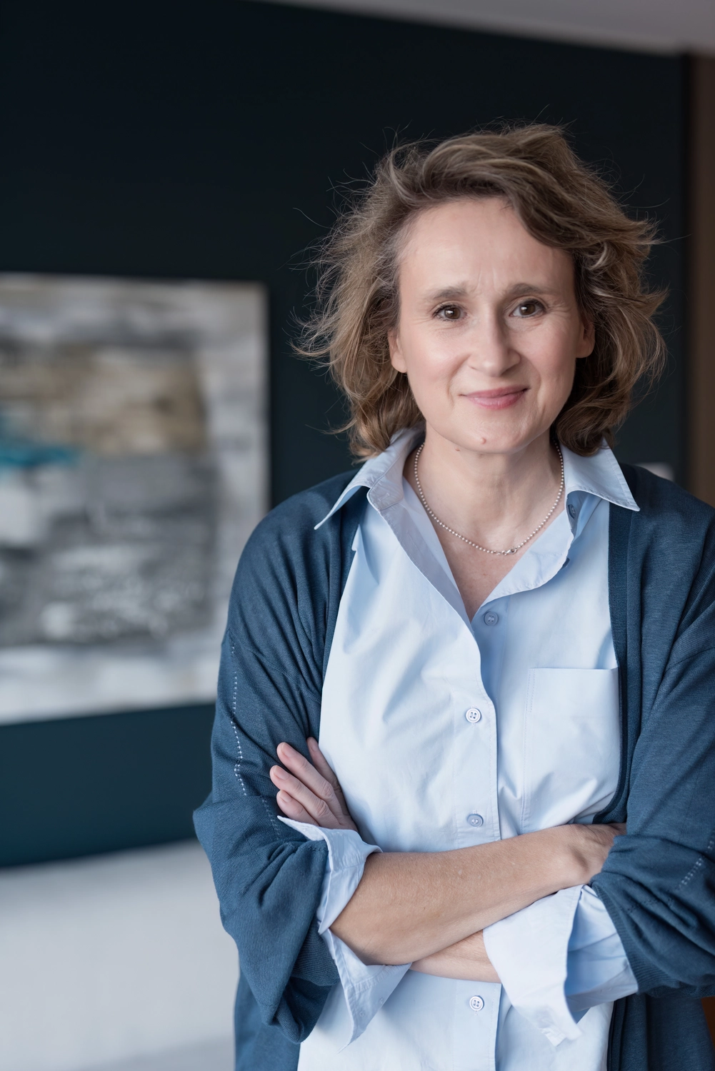 Portrait1 frontal Gesicht und Oberkörper Christiane Schicker vor einer Wand in dunkelblauem Anstrich und der Andeutung eines Wandbildes. CS trägt einen blauen Cardigan und eine hellblaue Hemdbluse