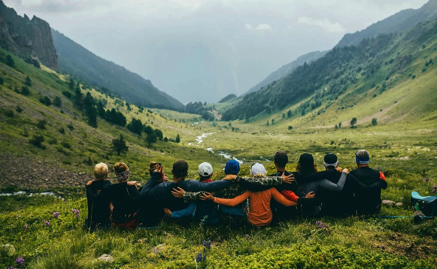 Blick von einem steilen grünen Berghang in ein wolkenverhangenes Tal. 11 Bergwanderer in Outdoorkleidung sitzen im Vordergrund am Hang in einer Reihe nebeneinander, Schulter an Schulter, sich mit den Armen gegenseitig Halt gebend. Sie blicken ins Tal runter, mit dem Rücken dem Fotografen zugewandt.
