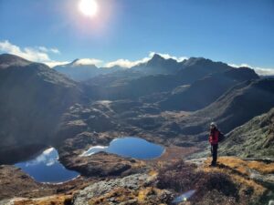 A hiker, Tashi Chozom, looks under a bright blue sky and in the backlight of the sun at a panorama of small glacier lakes, in which the blue sky is reflected. The silhouette of a mountain peak panorama frame the scenery in the background.